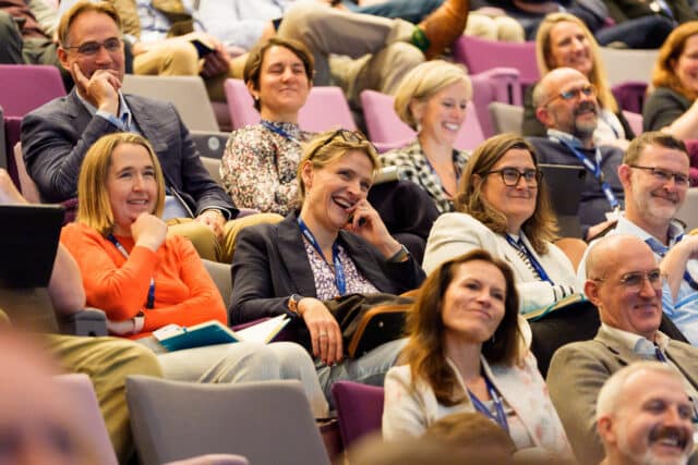 Audience members seated in a tiered conference auditorium, smiling and listening attentively during a presentation. Several attendees wear lanyards and hold notebooks or tablets.