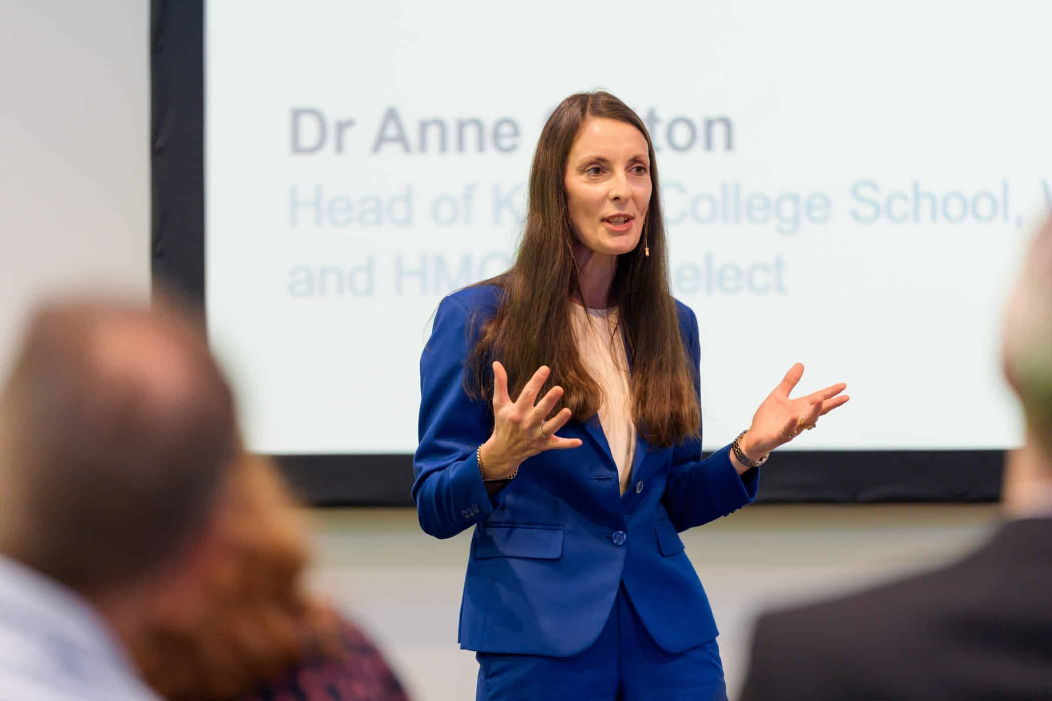 A woman in a blue suit speaks to an audience during a conference presentation, gesturing with her hands while standing in front of a projected screen displaying her name and title.