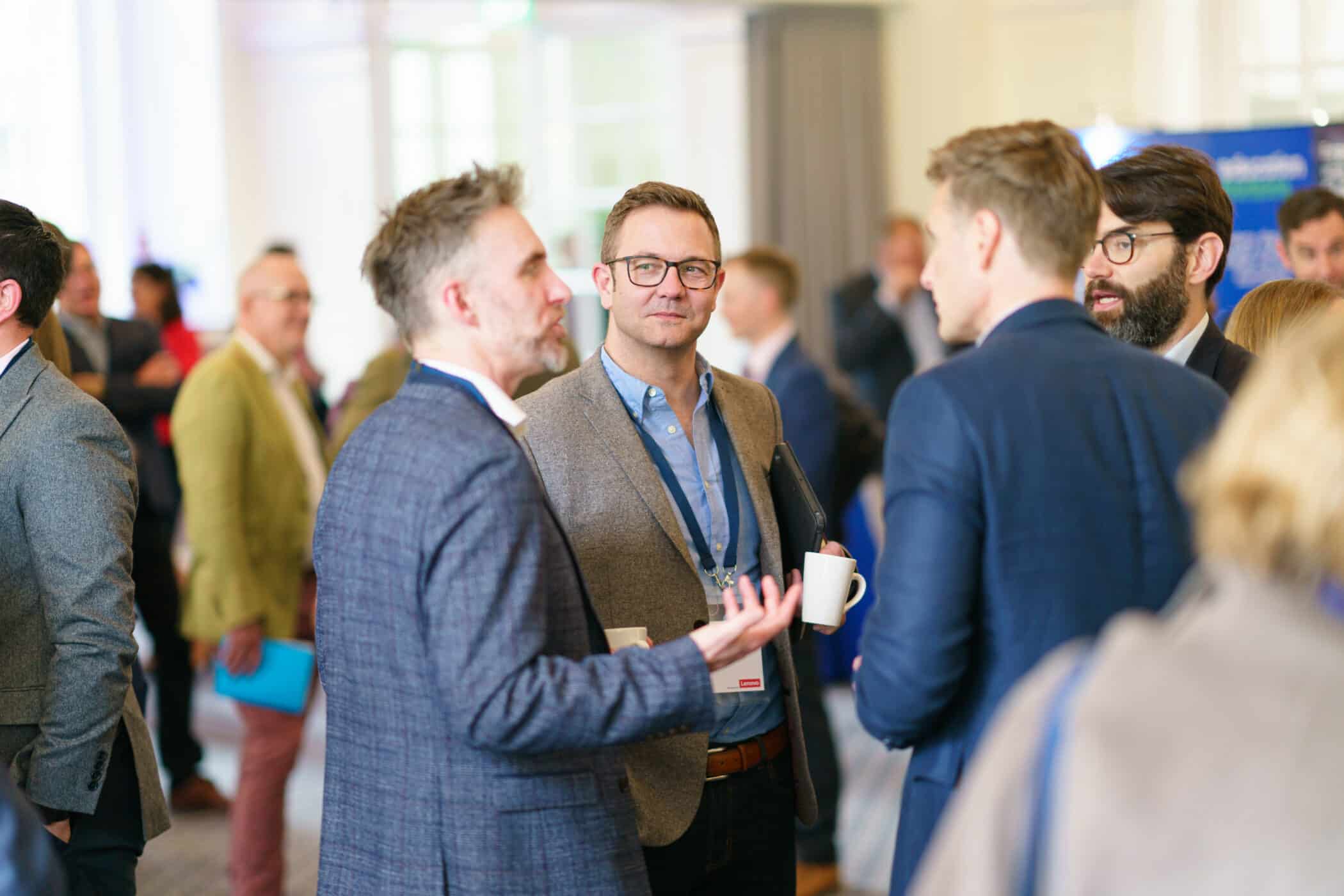 Attendees in business attire networking and conversing at a conference event, with several people holding coffee cups and wearing lanyards in a bright indoor venue.