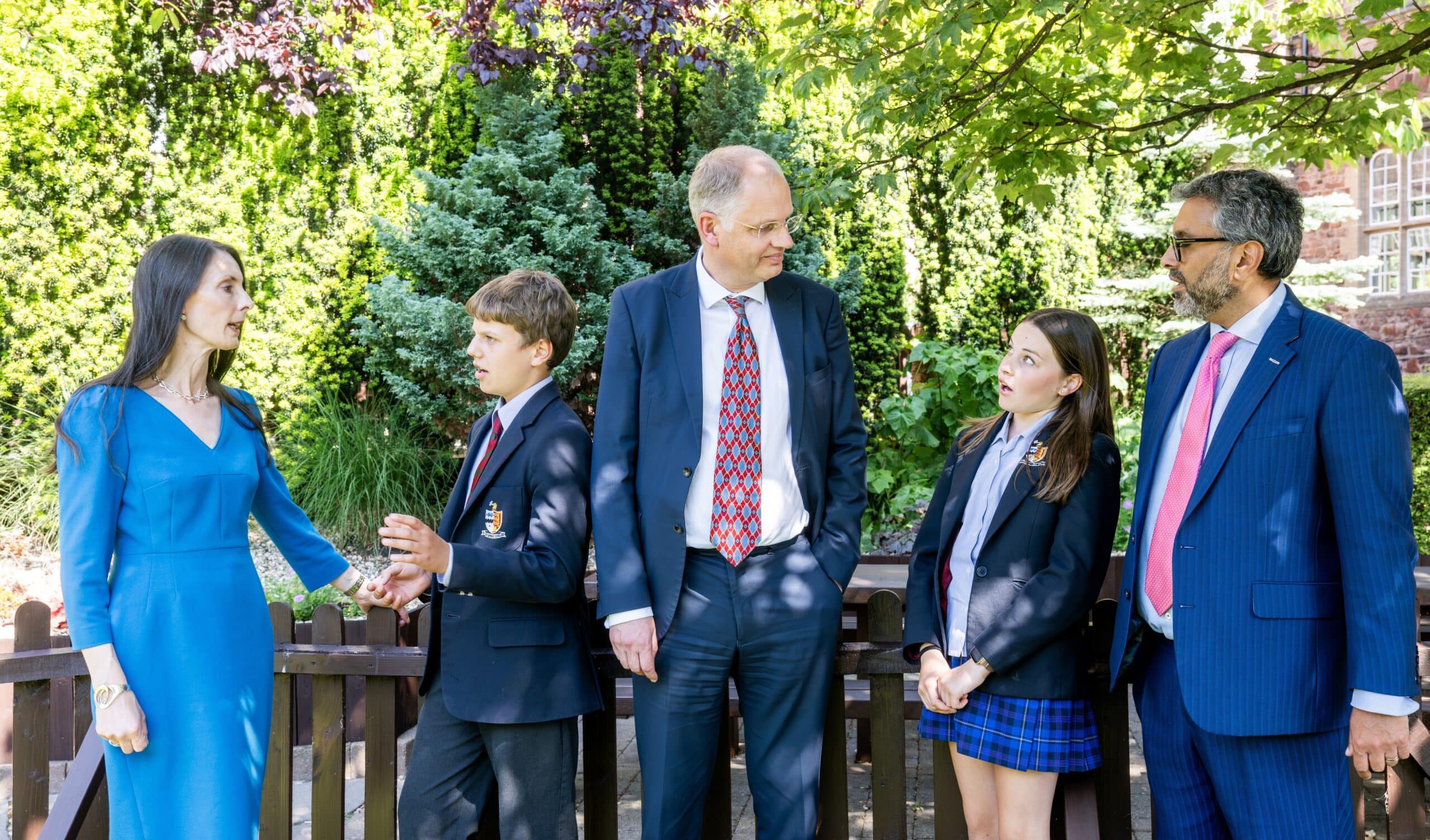 Five people stand outdoors in formal attire, including two adults in suits, a woman in a blue dress, and two students in school uniforms. They appear to be engaged in conversation, surrounded by lush greenery and a wooden fence.
