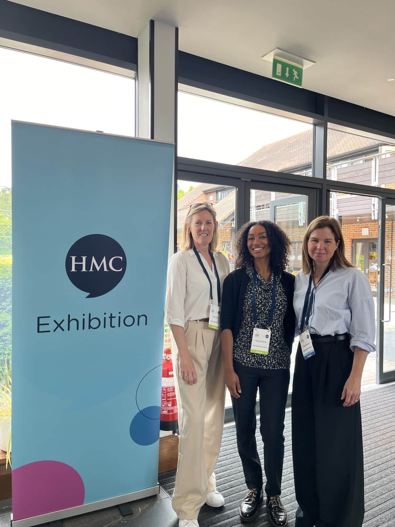Three women standing and smiling in front of a tall blue banner that reads “HMC Exhibition.” They are wearing lanyards and appear to be at a professional event inside a modern venue.