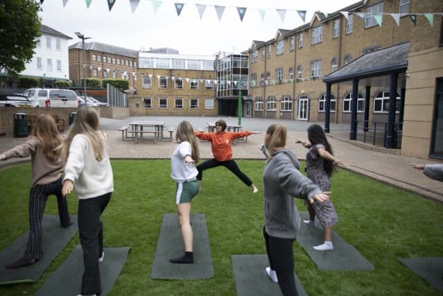 A group of students practicing yoga outdoors on a grassy area within a school courtyard. An instructor in an orange top leads the session, demonstrating a warrior pose, while students follow along on yoga mats. The setting includes surrounding school buildings and decorative bunting overhead.