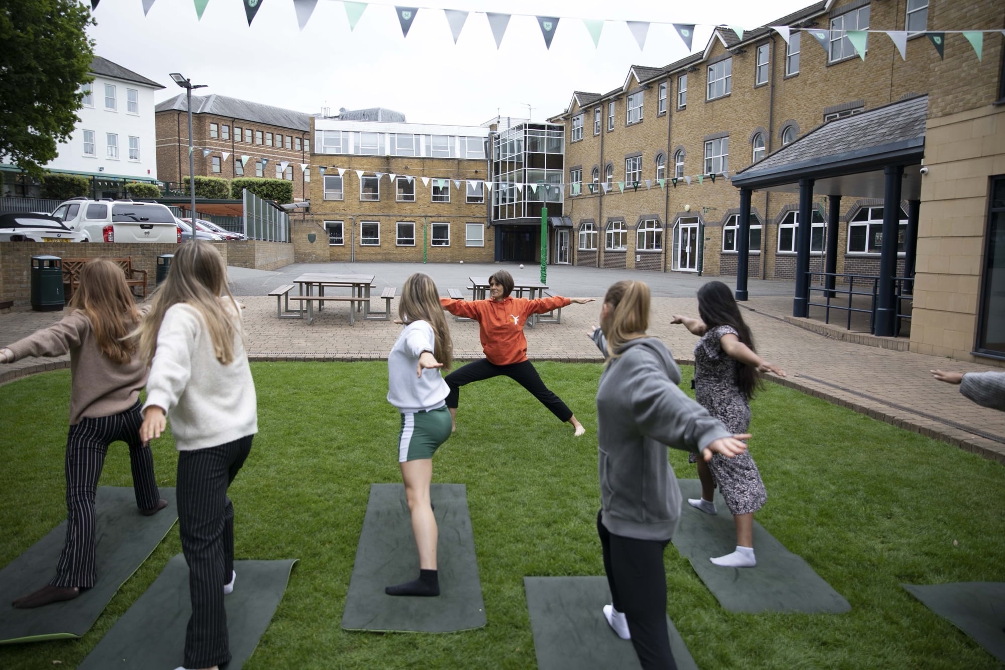 A group of students practicing yoga outdoors on a grassy area within a school courtyard. An instructor in an orange top leads the session, demonstrating a warrior pose, while students follow along on yoga mats. The setting includes surrounding school buildings and decorative bunting overhead.