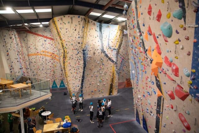 A wide view of a large indoor climbing centre with towering climbing walls covered in colourful holds. Several students wearing helmets and harnesses gather at the base of the wall, preparing for a climb. The facility includes tables, seating areas, and a visible mezzanine floor.