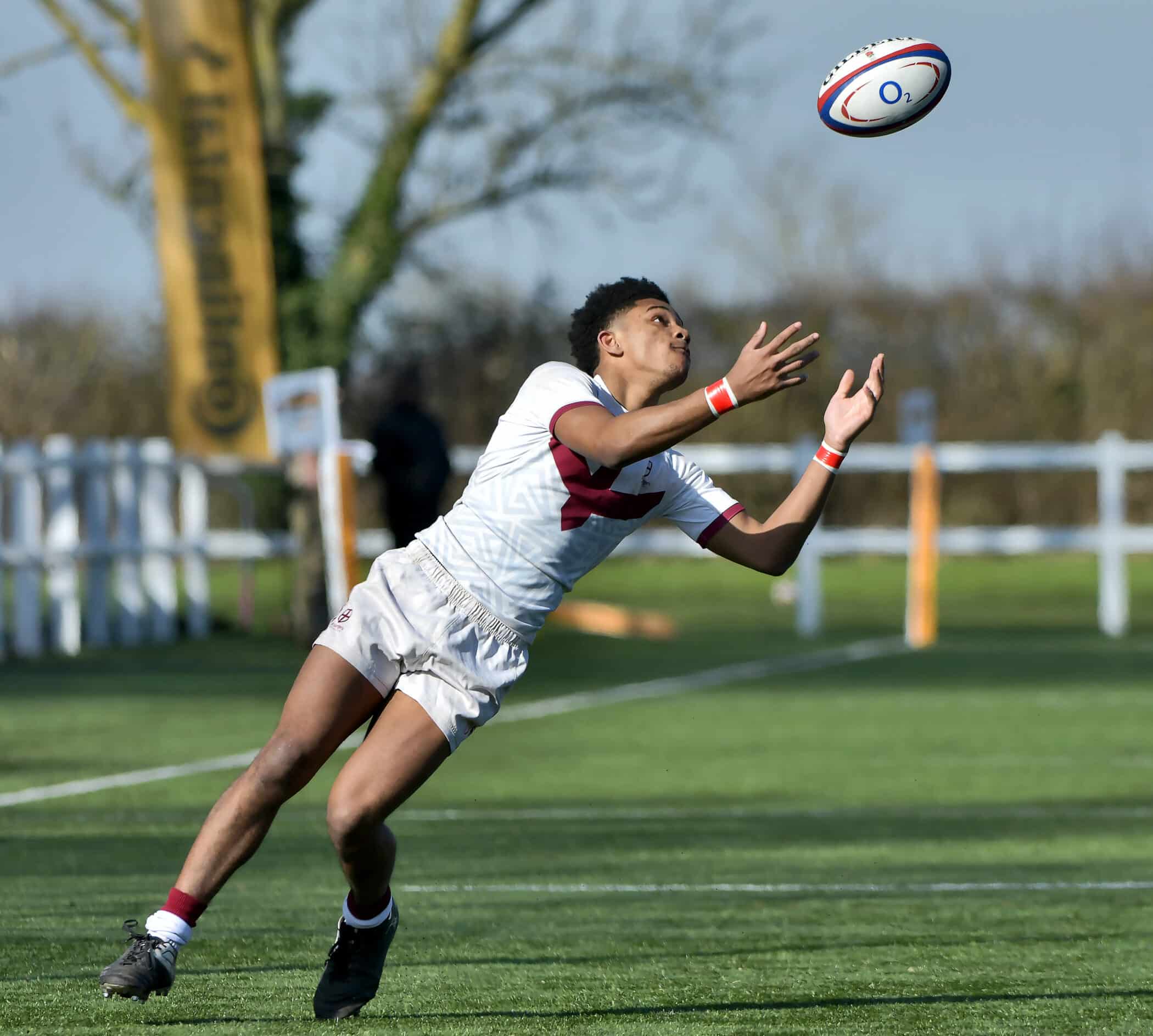 A young male rugby player dressed in a white and maroon uniform is mid-motion on a green pitch, reaching up to catch an incoming rugby ball under a sunny sky, with trees and fencing in the background.