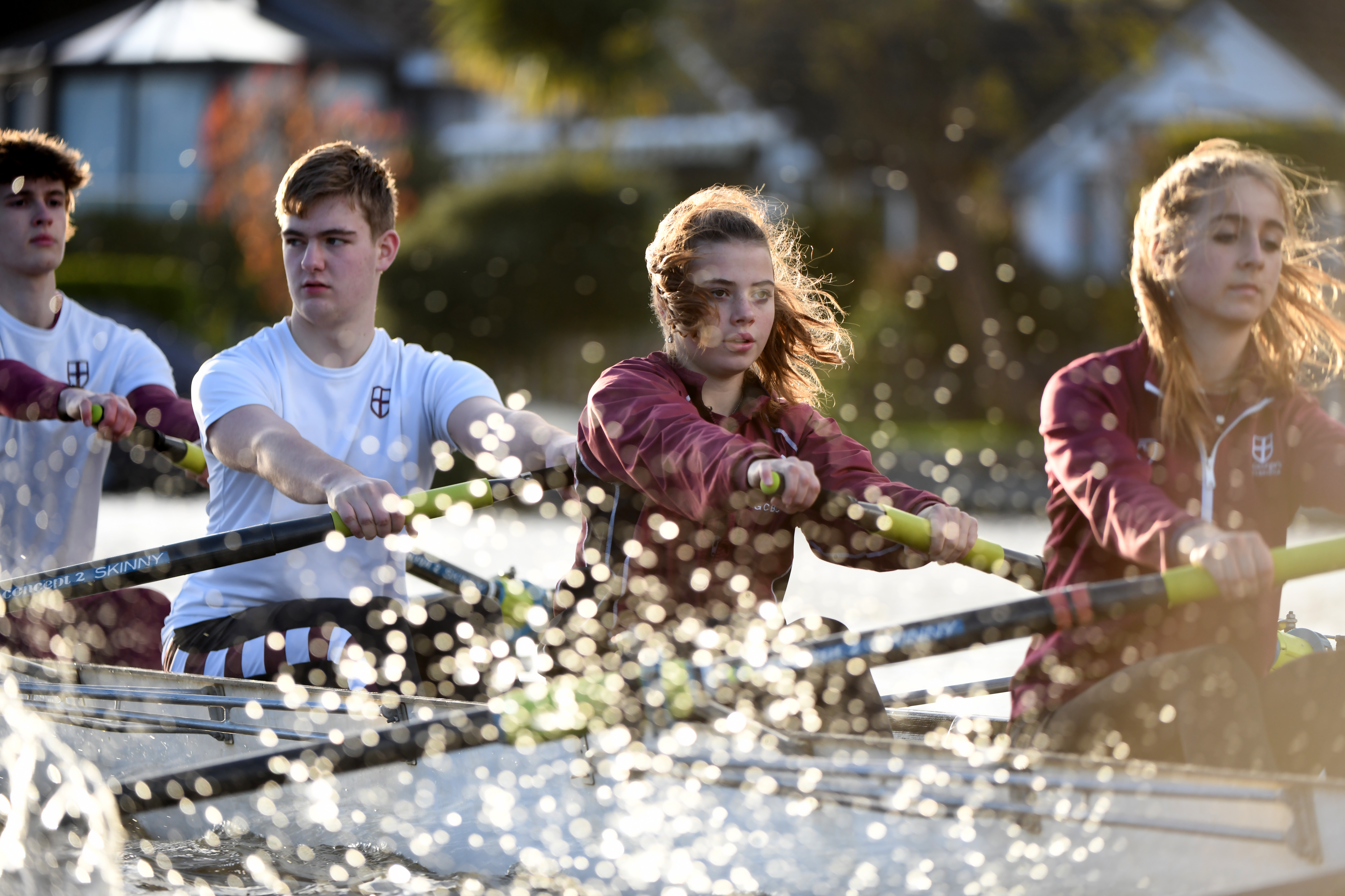 Mixed-gender students in maroon and white uniforms row together in a sculling boat on a river. Water splashes dramatically around them as they concentrate and pull in unison, with sunlight highlighting their determination.