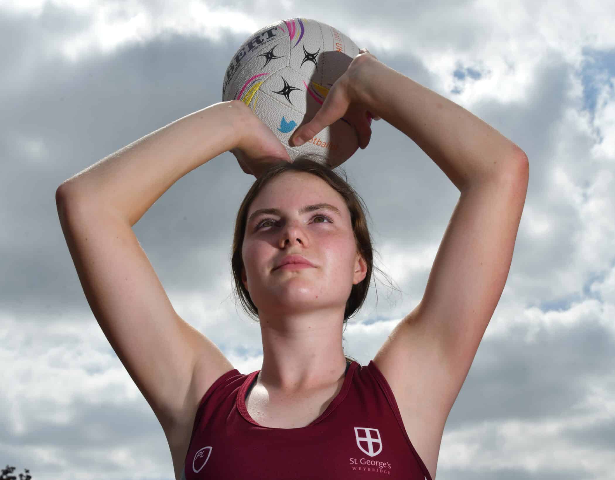 A young female netball player in a maroon St George’s Weybridge kit holds a netball above her head, preparing to pass or shoot, set against a cloudy outdoor sky backdrop.