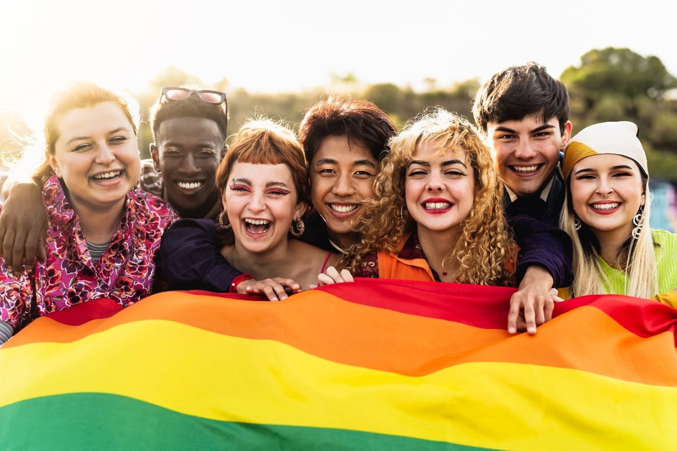 A diverse group of seven smiling young adults standing close together outdoors, holding a rainbow Pride flag. They appear joyful and united, symbolizing LGBTQ+ pride, inclusivity, and friendship.