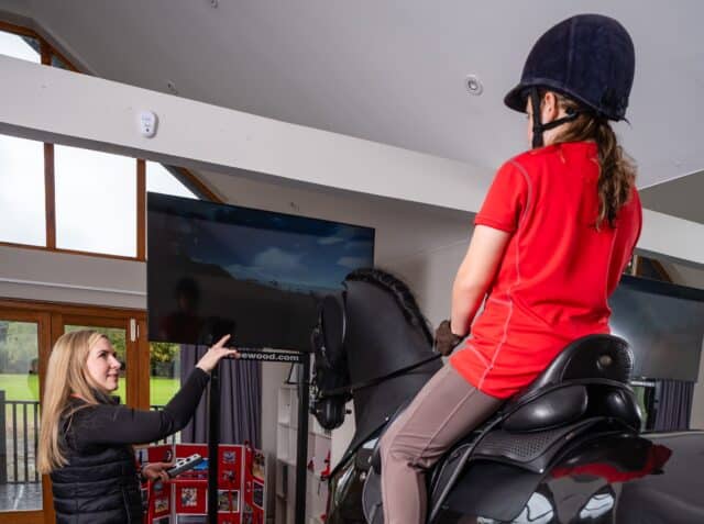 A young rider wearing a helmet and red shirt sits on a black equestrian simulator horse indoors. An instructor in black equestrian attire stands beside the simulator, gesturing and offering guidance. The room has wooden floors, a large screen displaying a riding simulation, and bright natural light coming through windows in the background.