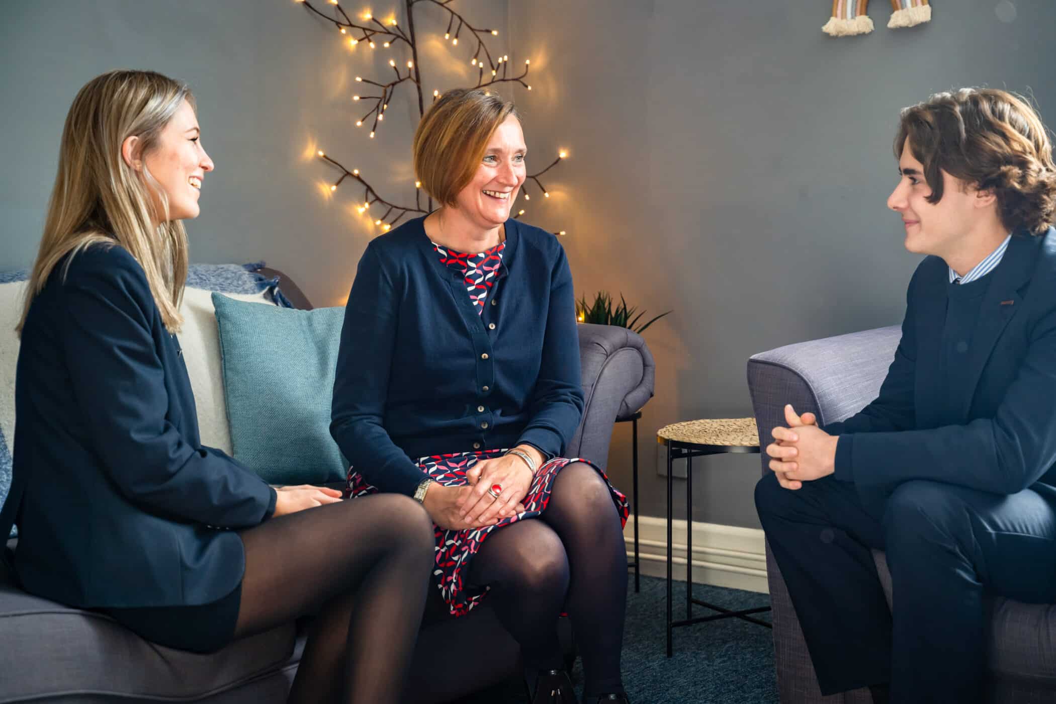 Two students in uniform sit on a sofa, smiling and talking with a woman in a patterned dress and navy cardigan. They are in a cozy, softly lit room with decorative string lights on the wall behind them.