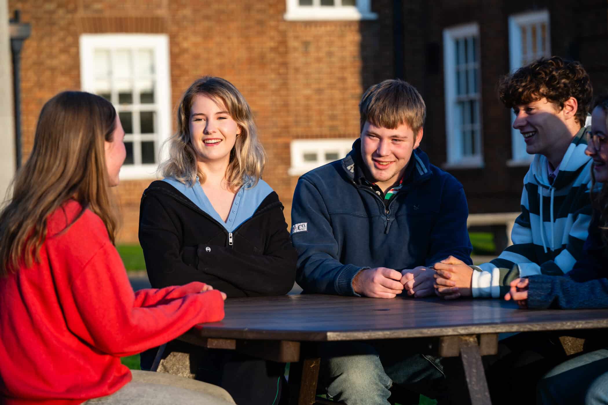 Five teenagers sit at an outdoor wooden table, laughing and talking in the sunshine. A red-brick school building is visible in the background, with white-framed windows catching the light.
