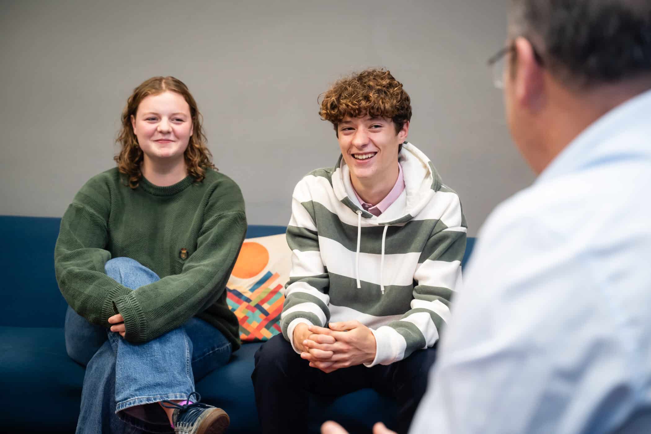 A teenage girl and boy sit on a blue sofa, smiling and talking to an adult whose back is to the camera. The setting is informal, with a grey wall and a colourful cushion in the background.