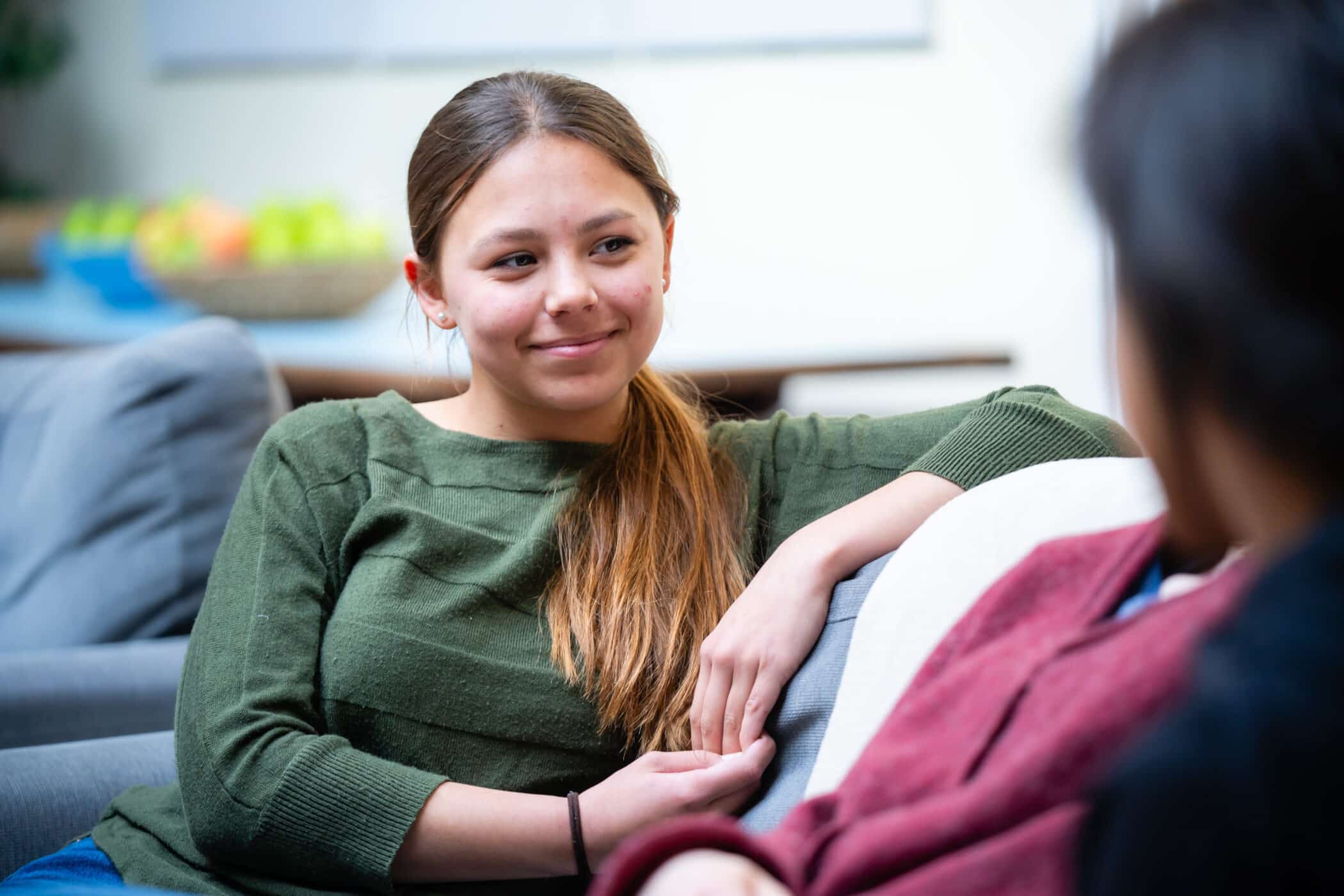 A teenage girl in a green sweater sits on a sofa, smiling warmly while listening to another person who is partially visible in the foreground. The setting is relaxed and informal, with a bowl of fruit blurred in the background.
