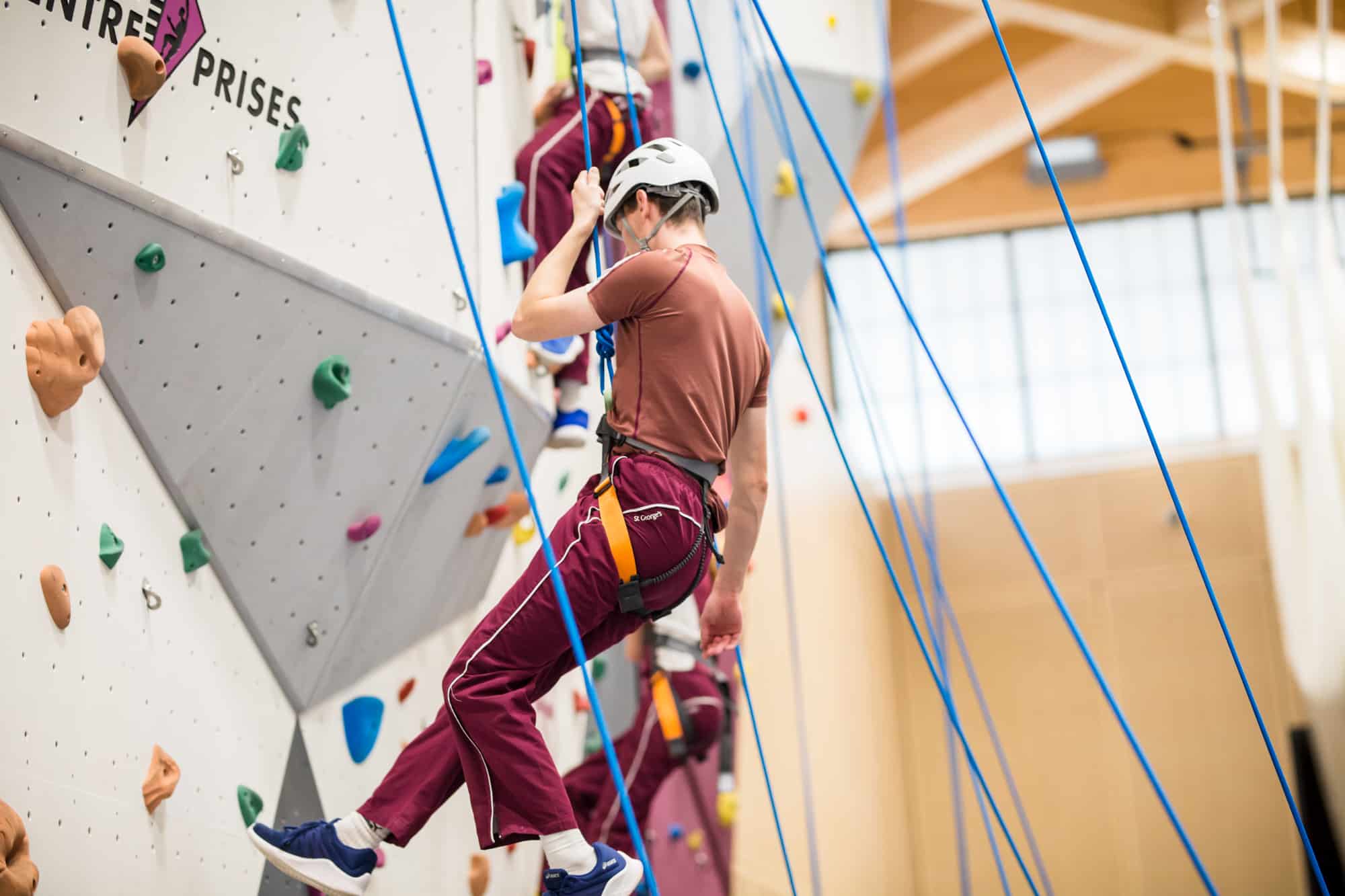 A student wearing a helmet, harness, and maroon tracksuit climbs a brightly coloured indoor rock wall with blue safety ropes, captured mid-ascent in a well-lit climbing gym.