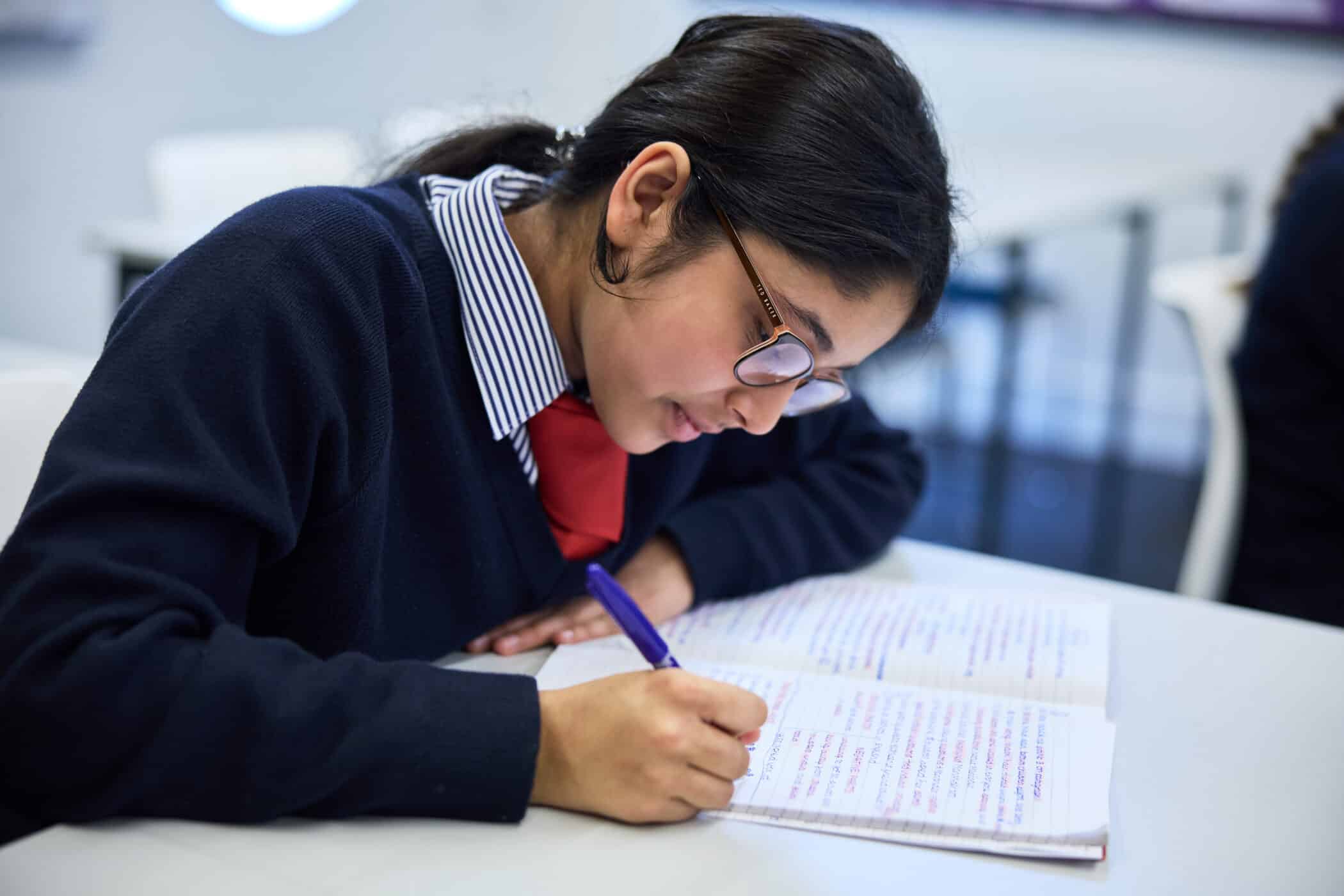 A female student in a navy sweater, red tie, and glasses is focused on writing notes in a lined notebook with a purple pen. She is seated at a desk in a classroom setting.
