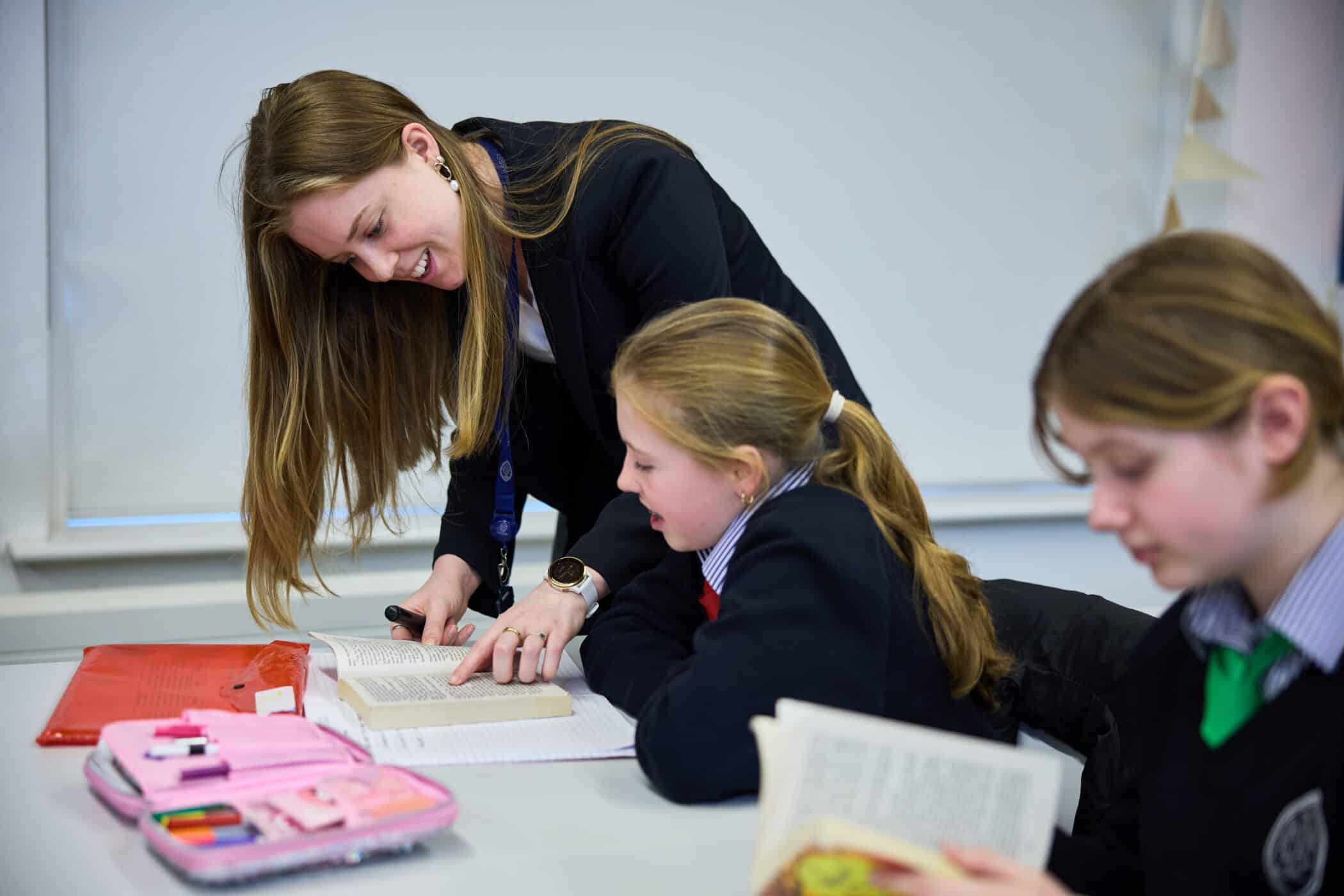 A teacher leans over to help a young student with reading, pointing at a passage in an open book. Two students in school uniforms are seated at a desk with books and pencil cases. One student in the foreground is reading independently.