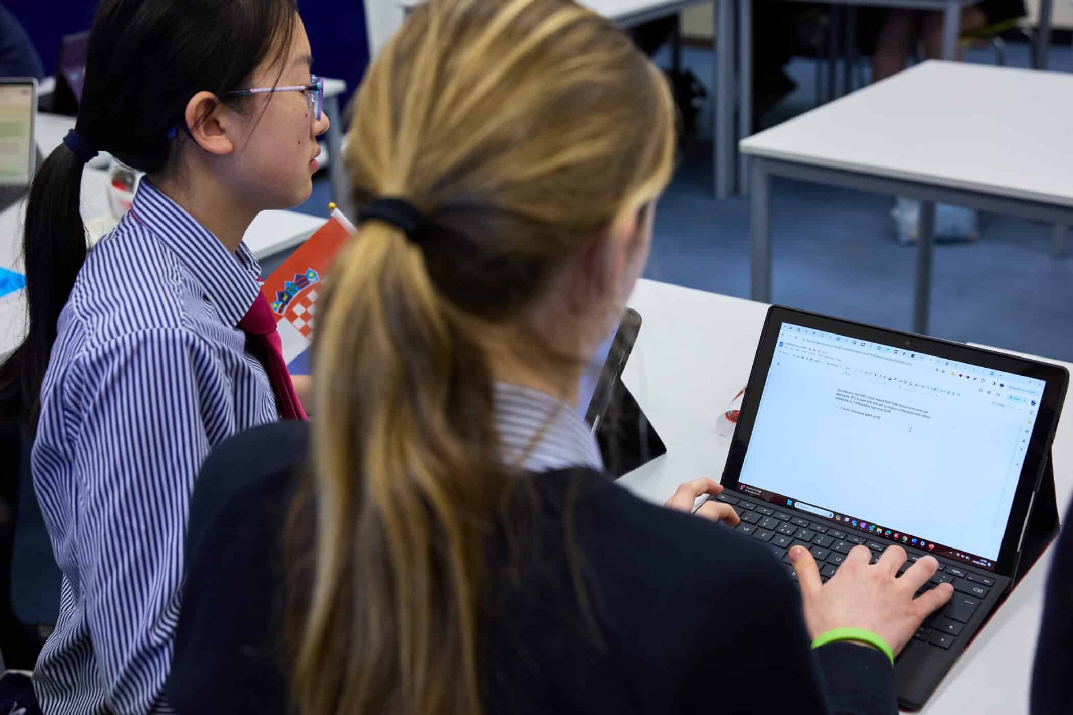 Two female students in school uniforms are seated at desks in a classroom, working together. One student is typing on a laptop with a document open on the screen, while the other looks on attentively. The classroom has several white desks and a blue carpeted floor. A Croatian flag can be seen partially in the background, suggesting an international or cultural context.
