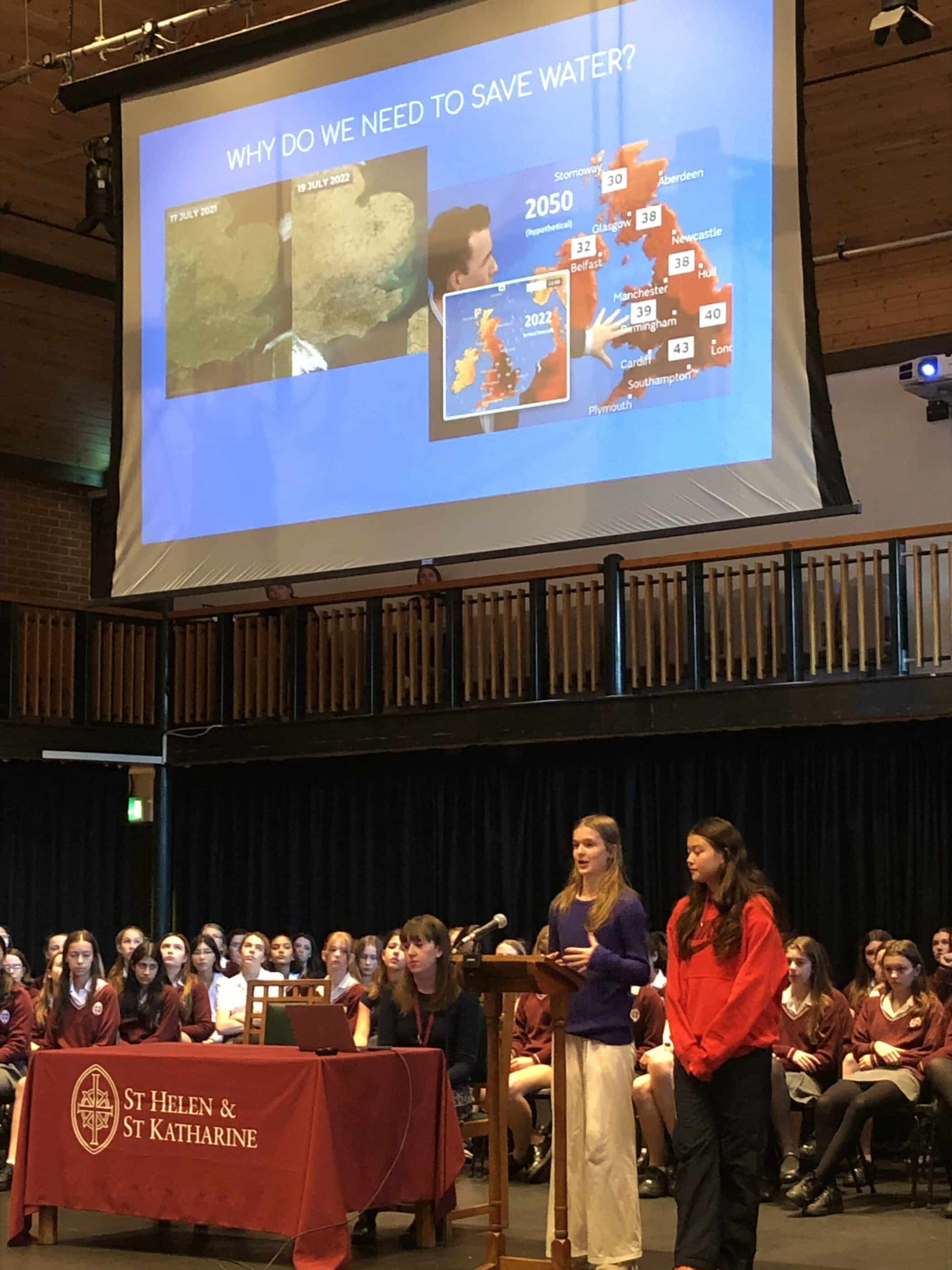 School assembly at St Helen & St Katharine on water conservation, with slides showing weather maps, future temperature projections, and students presenting on stage.