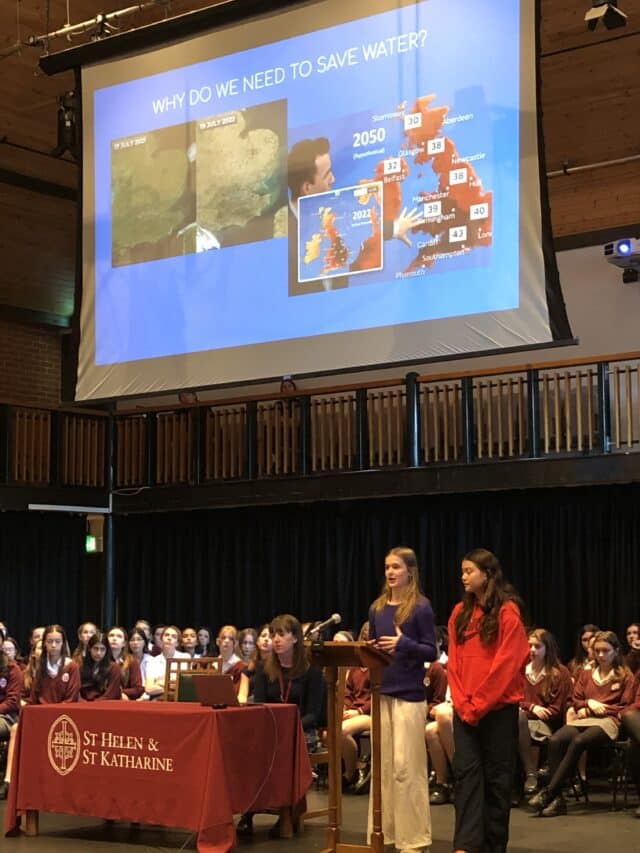 School assembly at St Helen & St Katharine on water conservation, with slides showing weather maps, future temperature projections, and students presenting on stage.