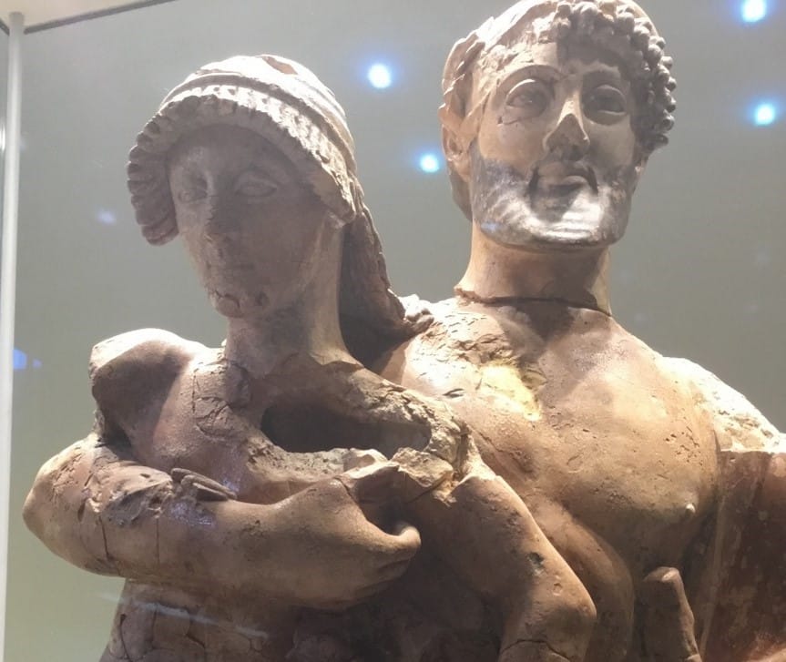 A weathered terracotta sculpture of Zeus and Ganymede, both draped in robes. The man holds the woman closely, and she cradles a small child or animal figure. The statue shows signs of damage and age, with visible cracks and missing fragments, suggesting antiquity. Displayed in a glass museum case under spotlighting.