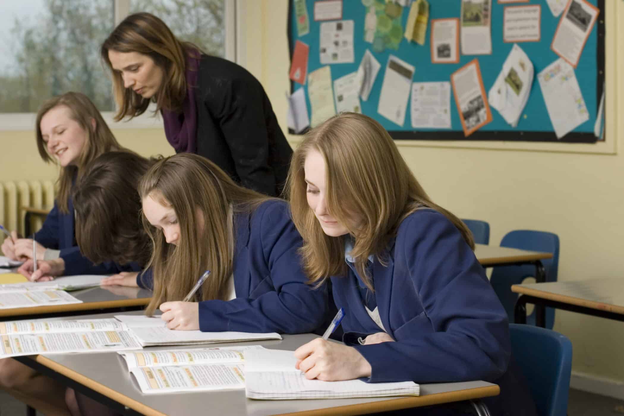 A teacher leans over a group of four secondary school girls in navy blue uniforms who are seated at desks in a classroom. The students are focused on writing in notebooks and referring to textbooks. Behind them, a bulletin board displays various educational materials and notices pinned on a bright blue background.