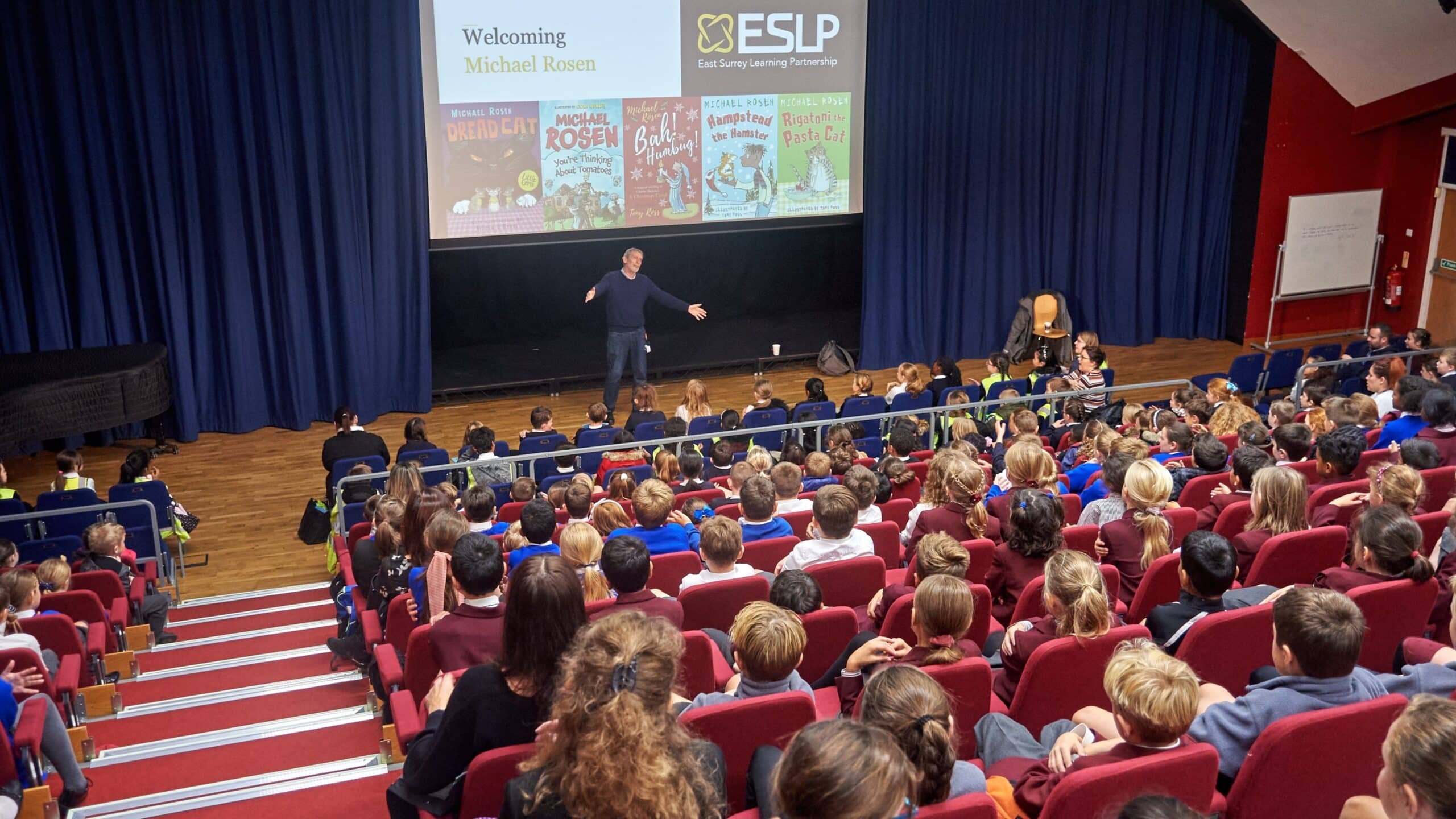 Author Michael Rosen stands on stage speaking to a large group of school children seated in an auditorium. A presentation slide behind him displays the East Surrey Learning Partnership logo, a welcome message, and covers of several of his children's books.