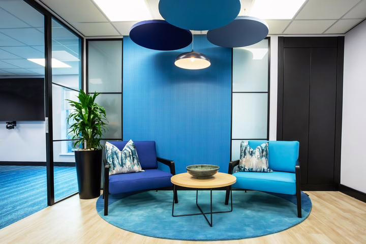 Modern lounge area with two blue armchairs and a round wooden coffee table on a blue circular rug. The space features a vibrant blue accent wall, decorative ceiling discs in varying shades of blue, and a pendant light. A tall green plant in a black pot adds a natural touch.