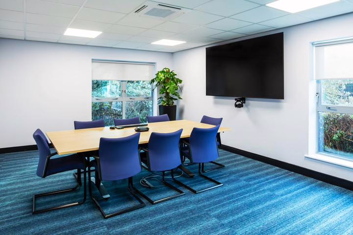 Meeting room with a large rectangular wooden table surrounded by blue chairs, equipped with a wall-mounted display screen and video conferencing equipment. Bright and professional interior with natural lighting.