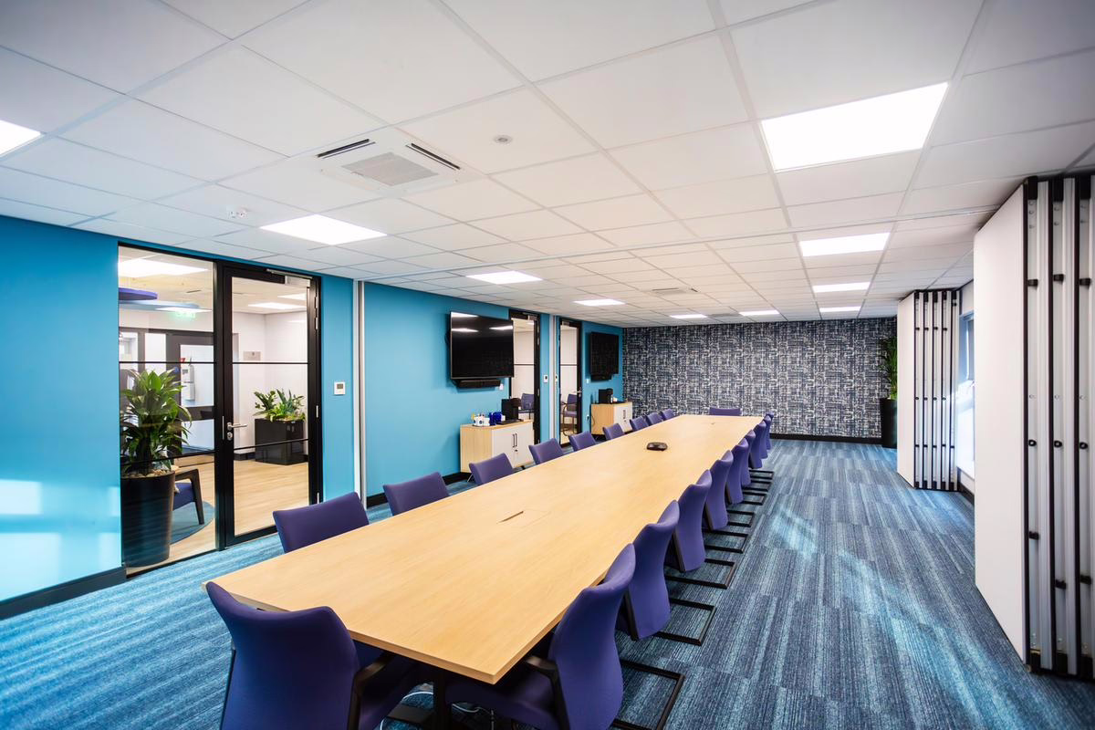 Spacious boardroom with a long central conference table and multiple blue chairs. The room includes glass walls, plants, and mounted AV equipment, offering a clean, functional layout.