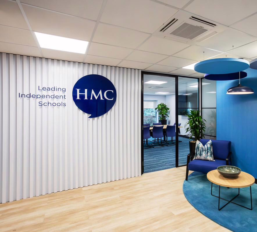 Reception area at HMC headquarters with a blue logo on a white slatted wall, stylish blue armchairs, indoor plants, and visible glass-walled meeting rooms in the background.