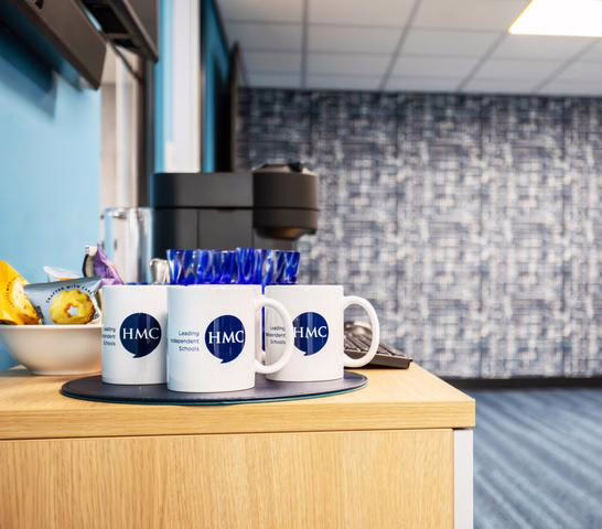 Refreshment station with a wooden round table holding several branded HMC mugs, positioned against a textured circular wall feature.
