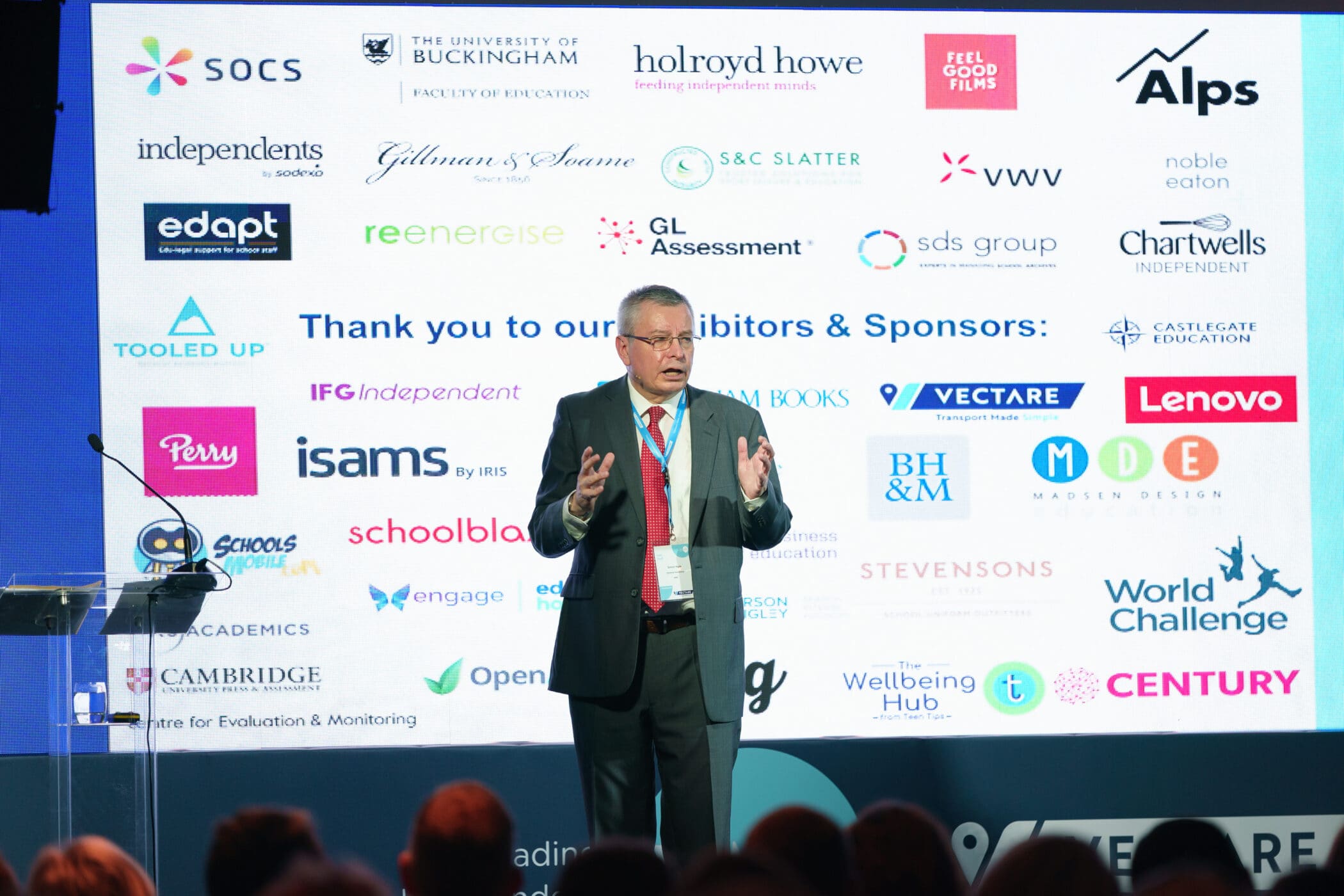 A man in a business suit speaks on stage in front of a large screen displaying logos of exhibitors and sponsors at an educational or professional conference. The audience is partially visible in the foreground.