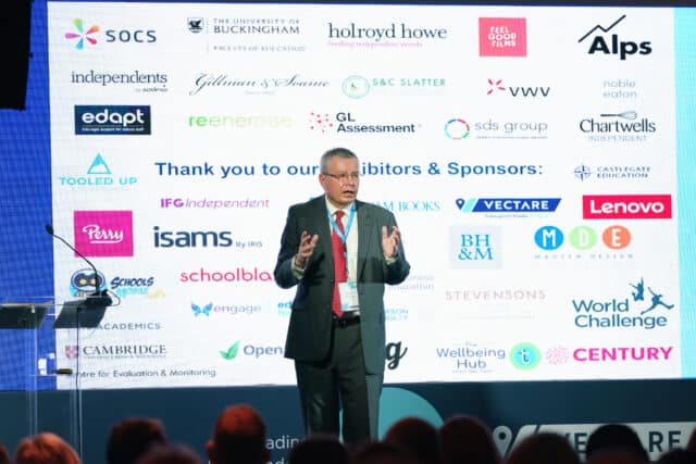 A man in a business suit speaks on stage in front of a large screen displaying logos of exhibitors and sponsors at an educational or professional conference. The audience is partially visible in the foreground.
