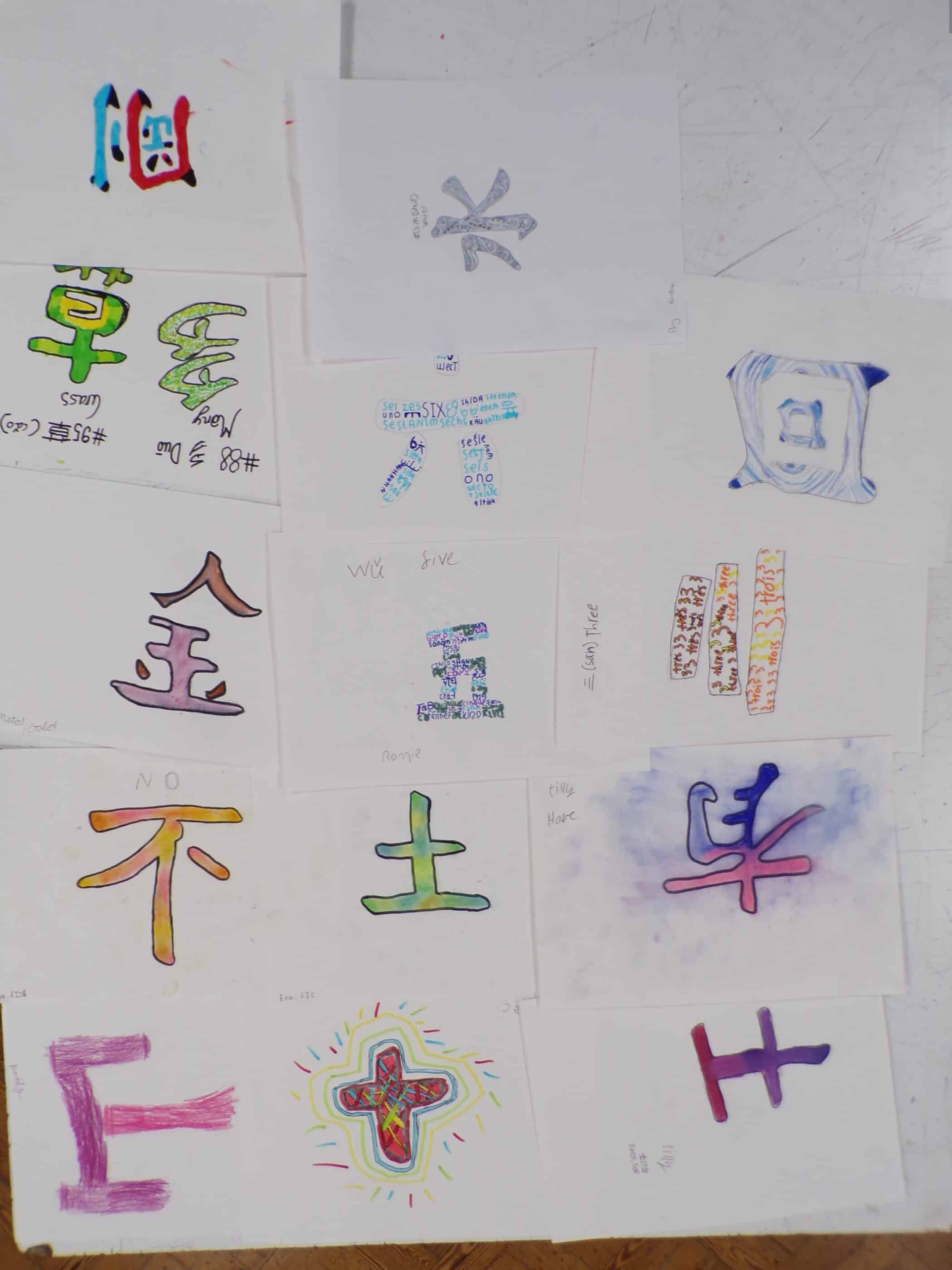 A collection of hand-drawn artworks on white paper, each featuring different colorful interpretations of Chinese or Japanese characters. The characters are drawn in various styles using markers, watercolors, and colored pencils, with some decorated using patterns, textures, or words. The sheets are spread across a wooden floor, arranged in a loose grid.