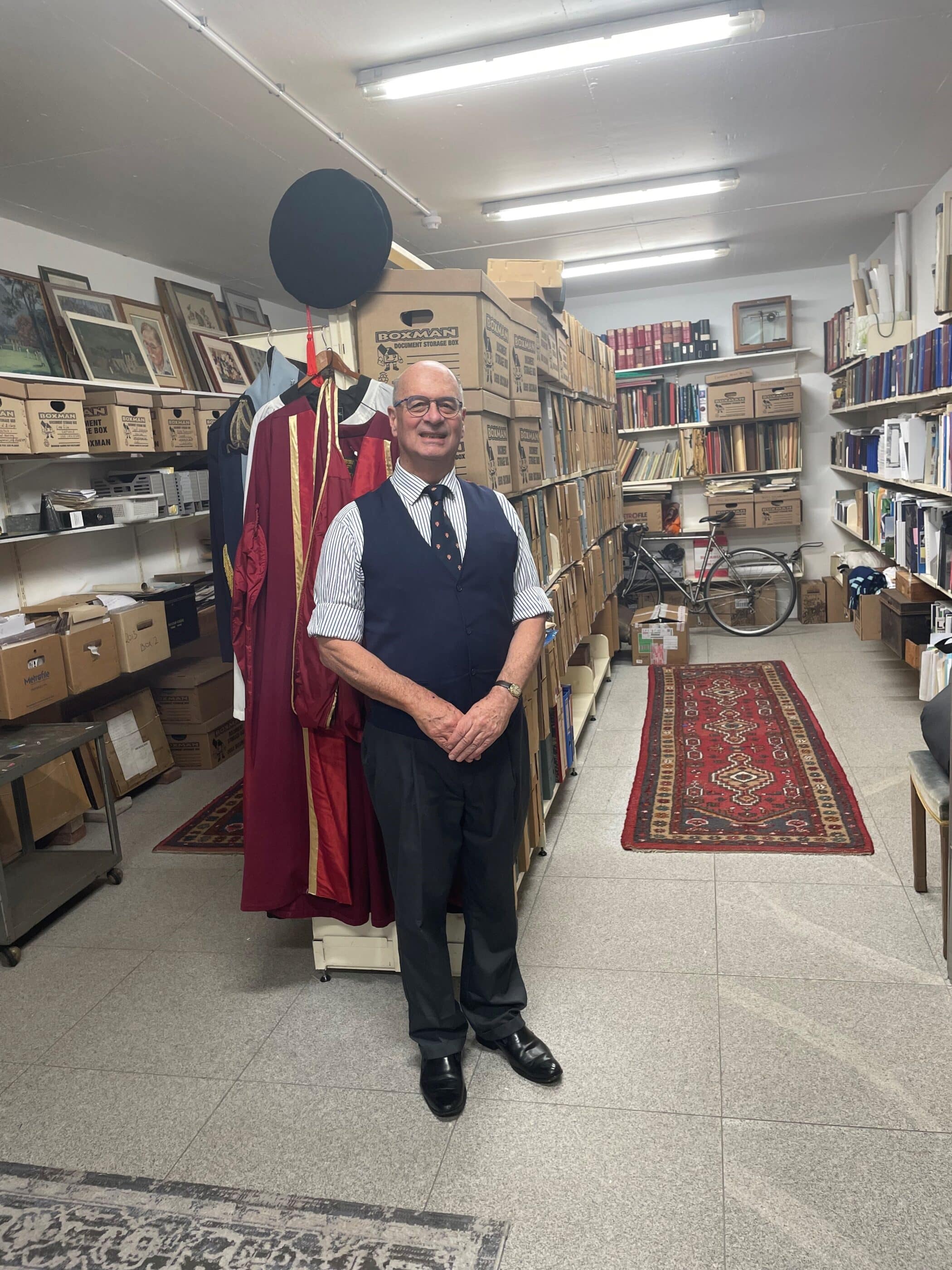 A smiling man in formal attire stands in a well-organized archive room filled with labeled storage boxes, bookshelves, and framed pictures. Behind him are ceremonial academic robes and a bicycle; the floor is lined with patterned rugs.