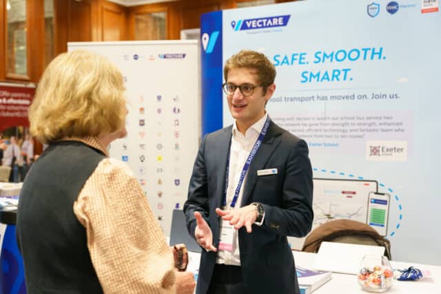 A man wearing a navy suit and exhibitor badge speaks with a woman at a trade show booth for Vectare, a school transport service. Behind him is a branded display with the slogan “Safe. Smooth. Smart.” and promotional materials on the table.