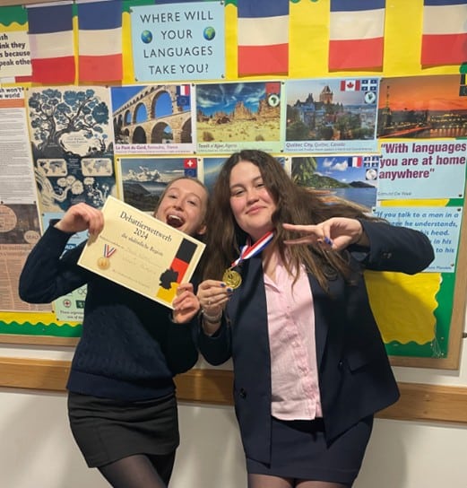Two secondary school students standing in front of a colorful bulletin board with world languages posters. One holds a certificate, the other a medal, both smiling and posing playfully for the camera.