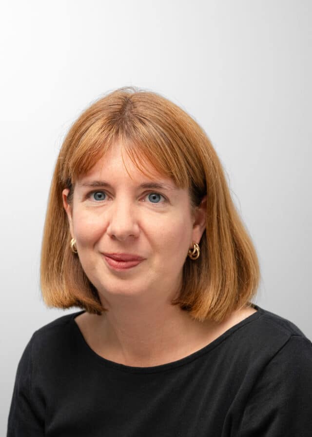Professional head-and-shoulders portrait of a woman with shoulder-length red hair and gold hoop earrings, wearing a black top against a light grey background.