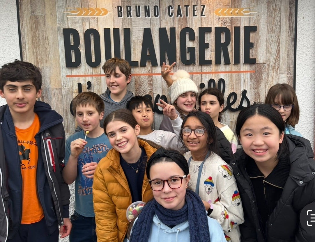 A group of young students standing outside a bakery called “Bruno Catez Boulangerie.” They are smiling, bundled in coats, and holding sweets or lollipops.