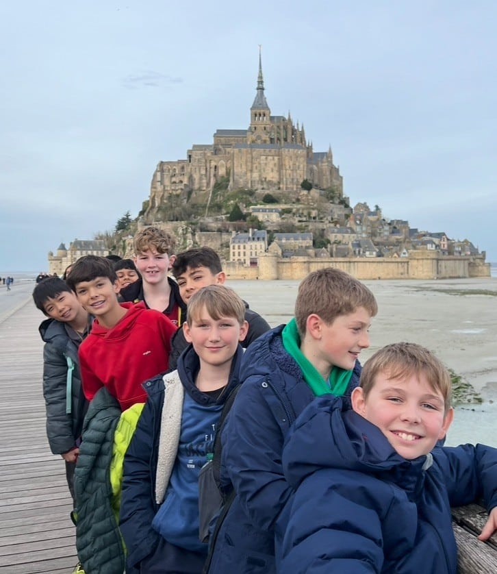 A group of boys standing on a wooden bridge with Mont Saint-Michel, a historic abbey in France, in the background. They are smiling and posing together.