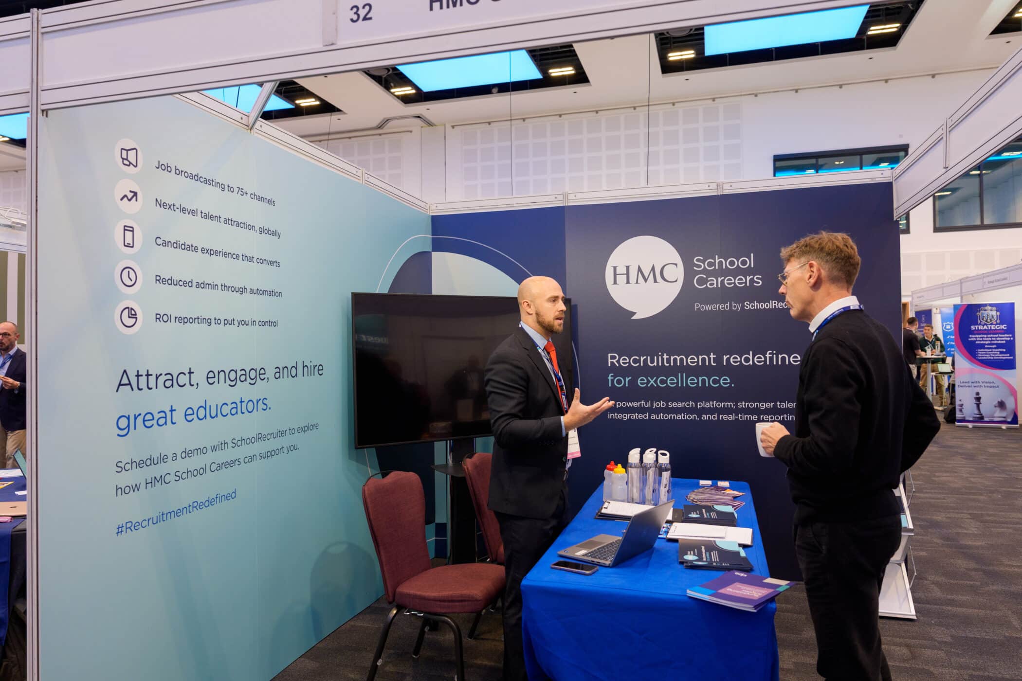 Two men talk at an exhibition booth for HMC School Careers, featuring branding and promotional text on the walls. One man in a suit gestures while speaking; the other listens holding a cup. The booth promotes recruitment services for educators, with brochures, water bottles, and a laptop on the table.