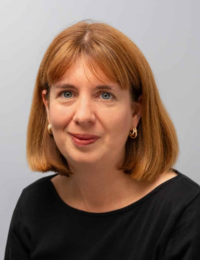 A woman with shoulder-length auburn hair and a fringe, wearing a black top and gold hoop earrings. She has a slight smile and faces the camera against a plain light grey background.