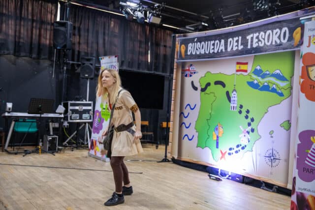 A woman dressed in a safari-style beige costume with a belt and crossbody bag is performing on stage. Behind her is a large illustrated backdrop of Spain and Portugal labeled “Búsqueda del Tesoro” (Treasure Hunt), decorated with symbols like mountains, a windmill, and footprints. Stage equipment, speakers, and banners for “Joy 4 Kids” are visible around the set.
