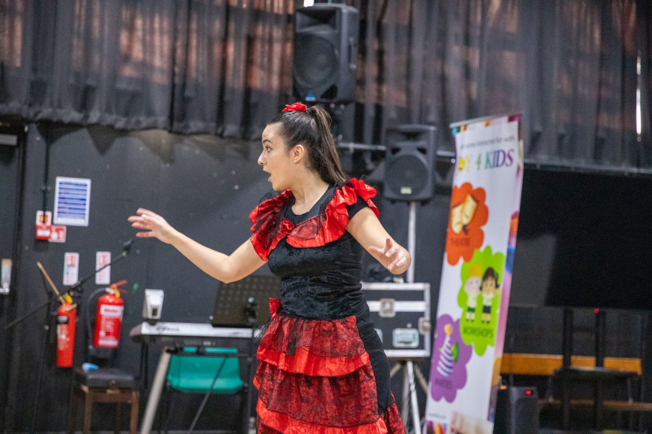 A woman dressed in a red and black flamenco-style costume, with layered ruffles and a red hair accessory, performs animatedly on stage. She has her arms extended and an expressive face. Behind her are banners for “Joy 4 Kids” and stage equipment, including speakers and musical instruments.