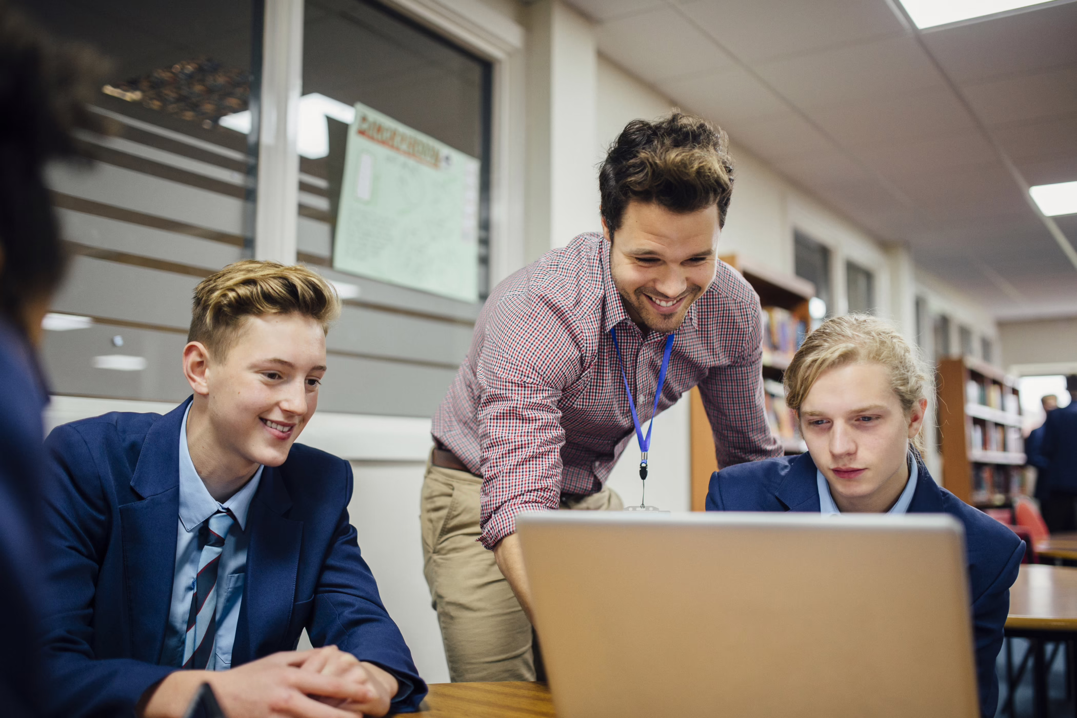 A smiling male teacher stands beside two students in school uniforms, looking at a laptop together in a classroom or library setting.