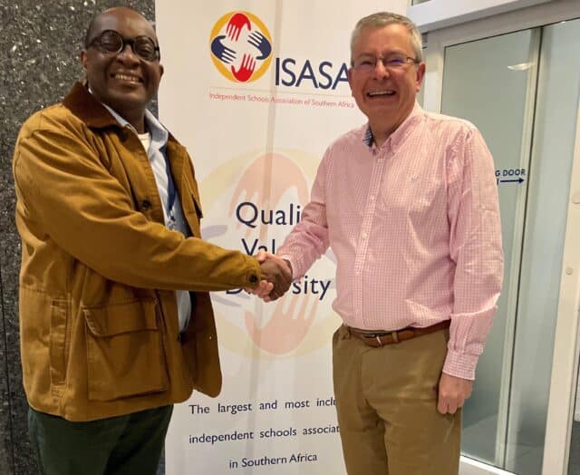 Two men smiling and shaking hands in front of a banner for ISASA (Independent Schools Association of Southern Africa). The banner features the organisation's logo and slogan promoting quality, values, and diversity.