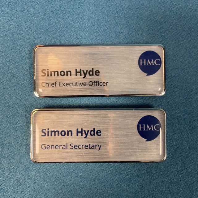 Two rectangular metallic name badges on a blue surface, both labelled "Simon Hyde" with the HMC logo. The top badge reads "Chief Executive Officer" and the bottom one reads "General Secretary."