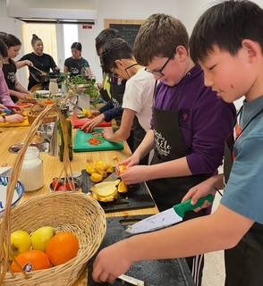 A group of students wearing aprons preparing fruit at a long table during a cooking class. Several are slicing oranges and other ingredients.