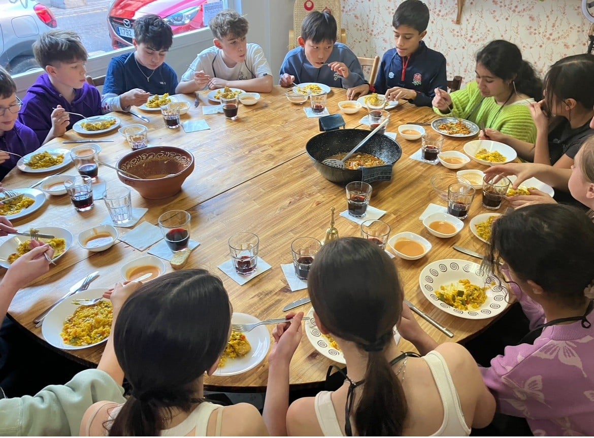 A group of students sitting around a large wooden table, enjoying a shared meal together. Plates of food, glasses of juice, and bowls are set across the table.