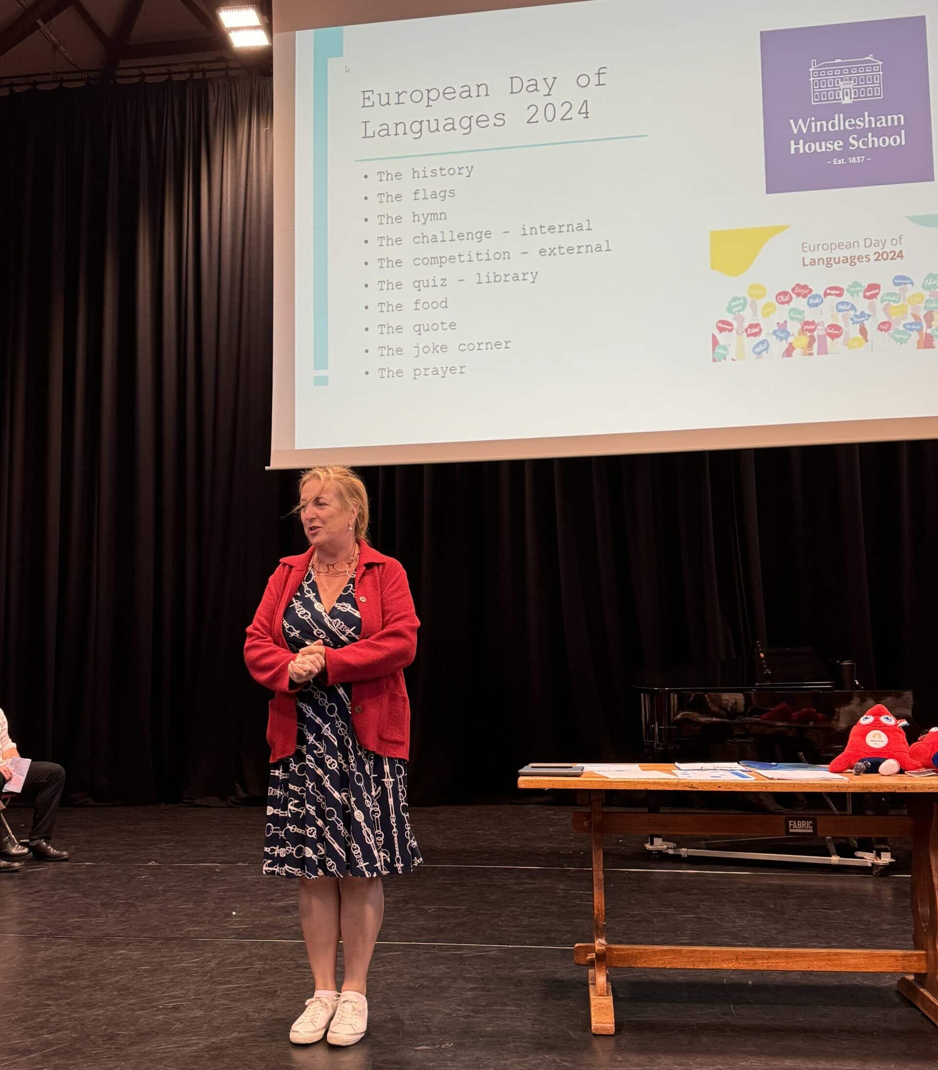A woman in a navy patterned dress with a red cardigan stands on stage addressing an audience. Behind her, a large projector screen displays a presentation slide titled “European Day of Languages 2024” with a bullet point agenda, alongside the Windlesham House School logo.